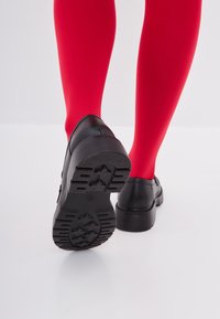 Czarne buty z teksturowaną podeszwą, okrągłym noskiem i minimalnymi zdobieniami, w połączeniu z jasnoczerwonymi rajstopami. Prosty, funkcjonalny design.