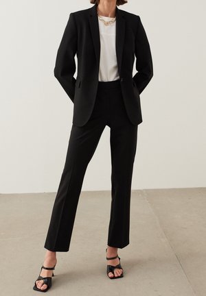 Blazer noir ajusté et pantalon assorti, associé à une chemise blanche. Présente une coupe slim, un tissu lisse et des sandales à talons ouverts noires.