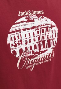 Camiseta de algodón burdeos con un gráfico blanco de un edificio y el texto "Jack & Jones Originals desde mil novecientos noventa."