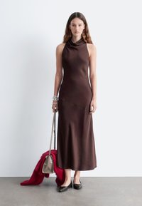 Braunes ärmelloses Kleid mit einem Cowl-Neck, glatter Textur und einer fließenden Silhouette. Begleitet von einem roten Schal und einer kleinen beigefarbenen Tasche.