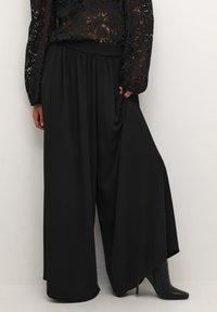 Pantalons larges noirs en tissu doux avec une taille élastique, présentant un design fluide et une texture lisse. Associés à des talons noirs.