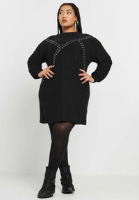 Abito maglione oversize nero con scollatura arrotondata, maniche lunghe e dettagli in borchie argentate; abbinato a collant neri e stivaletti alla caviglia.