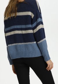 Kvinna iförd en stickad tröja med horisontella ränder i marinblått, ljusblått och krämvitt, i kombination med svarta byxor, sedd bakifrån.