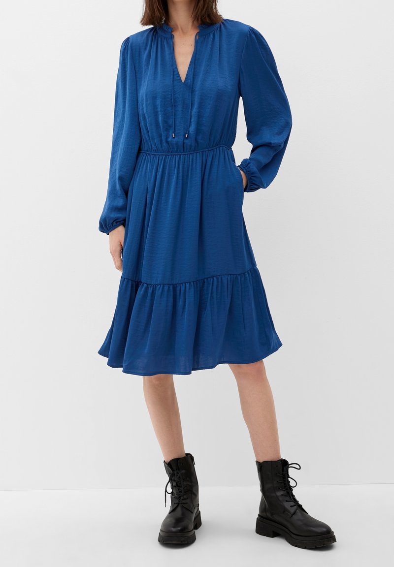 Femme portant une robe bleue à manches longues et longueur genou avec une taille froncée, associée à des bottes de combat noires à lacets, debout devant un fond uni.