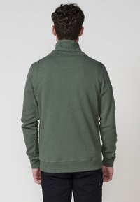 Hombre que lleva una chaqueta de forro polar verde oliva texturizada y pantalones negros, de pie de espaldas contra un fondo claro y liso.
