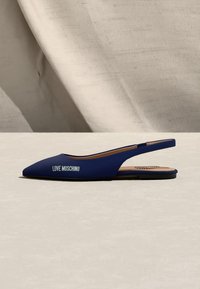 Πλατφόρμα slingback με μυτερή μύτη σε ναυτικό μπλε χρώμα, με λευκό λογότυπο "LOVE MOSCHINO" στο πλάι, τοποθετημένη σε μπεζ επιφάνεια με υφασμάτινο φόντο.