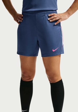 Personne portant un short de sport Nike bleu foncé avec un logo rose et des chaussettes noires montant jusqu'aux genoux, debout les mains sur les hanches.