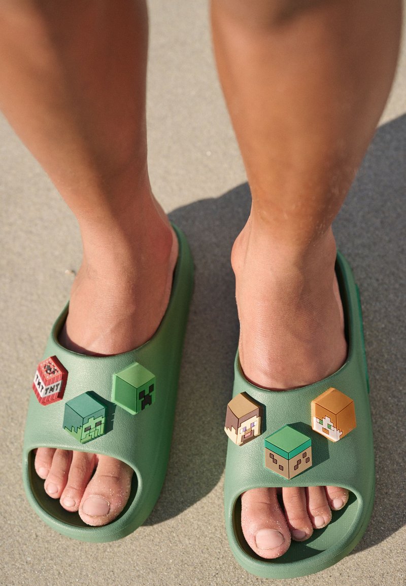 Sandales vertes à enfiler en matériau lisse, comportant quatre motifs de personnages Minecraft de différentes couleurs et formes sur le dessus.