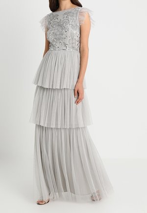 Kvinde iført en lysegrå, lagdelt tylkjole med udsmykket blomsterprydet overdel og gennemsigtige, flæsekantede kapærmer, stående foran en hvid baggrund.