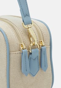 Beige strukturerad tygpåse med ljusblå läderdetaljer och guldfärgade dragkedjor. Har två dragkedjehandtag och en axelrem.