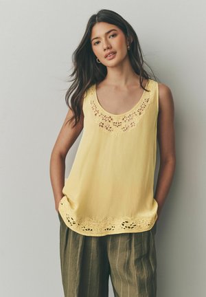 Blouse sans manches jaune avec des détails en dentelle délicats au niveau du col et de l'ourlet ; tissu doux qui tombe confortablement, associé à un pantalon olive à rayures.