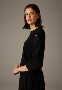 Pull noir en maille avec manches trois-quarts, présentant un motif floral texturé avec des accents de perles sur l'épaule gauche.