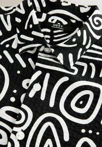 Svart t-shirt med korta ärmar och vita abstrakta mönster, knappad krage och plastknappar. Synlig etikett med "NEXT"-logotyp. Texturerad tyg.