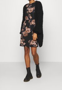 Czarna sukienka w kwiaty z różowymi i niebieskimi akcentami, zestawiona z fakturowanym czarnym kardiganem i czarnymi butami na sznurówki. Teksturowany splot i luźny krój.