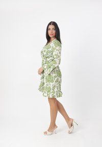 Vestido blanco con patrón floral verde, dobladillo con volantes y mangas largas. La modelo lleva zapatos de tacón alto y abiertos por delante. Fondo sencillo.