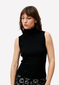 Zwarte geribbelde coltrui zonder mouwen, voorzien van subtiele glitters. Gestyled met zwart-grijze, patroonbroek.