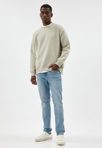 Felpa grigia chiara con polsini e orlo a costine, abbinata a jeans blu chiaro e sneaker bianche. Design semplice, vestibilità casual.