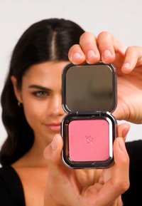 Kvadratisk kompakt blush i knallrosa med speil. Kompakten har en blank svart utside og en logo på undersiden.