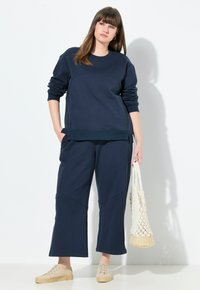 Marinblå sweatshirt och vida byxor set, tillverkat av mjukt tyg med geometriska sömmar. Kompletteras med en vävd väska och lätta skor.
