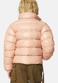 Giacca a collo alto di colore rosa, con sezioni trapuntate e una texture liscia e lucida. Il modello indossa pantaloni verde oliva.