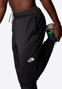 Black athletic pants with an elastic waistband, side pockets, and a white logo on the left thigh. Smooth fabric and tapered leg design.