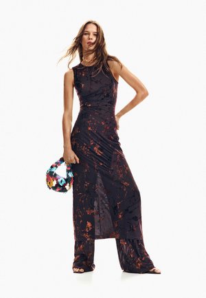 Mujer posando con vestido sin mangas negro y naranja con estampado sobre pantalones a juego, sosteniendo un bolso texturizado y colorido, con zapatos abiertos.