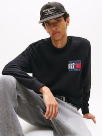 Černá mikina s modrým a červeným logem "tommy jeans" na hrudi, v kombinaci se světlými šedými džínami a černou kšiltovkou s bílým textem.