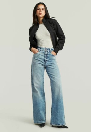 Jonge vrouw staat met handen in de zakken, draagt een zwart jasje met ritssluiting, lichtgrijs shirt, hooggetailleerde brede jeans en zwarte puntige schoenen.