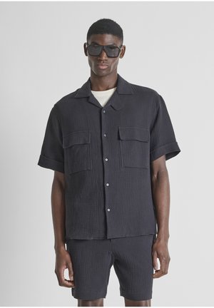 Man draagt een zwart getextureerd overhemd met korte mouwen en knopen, met twee borstzakken en bijpassende korte broek, gecombineerd met donkere oversized zonnebril.