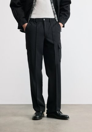 Osoba nosząca czarne spodnie typu cargo z przednimi plisami i kieszeniami, czarne eleganckie buty, białą koszulę oraz czarną kurtkę z kratkowaną podszewką.