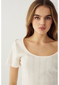 Junge Frau mit langen braunen Haaren, die ein cremefarbenes, kurzes Strickoberteil mit gemusterten vertikalen Linien und einem tiefen Rundhalsausschnitt trägt.