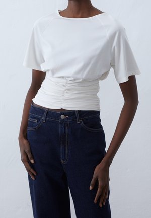 Persona che indossa una maglietta bianca arricciata a manica corta infilata in jeans blu scuro a vita alta su uno sfondo chiaro uniforme.