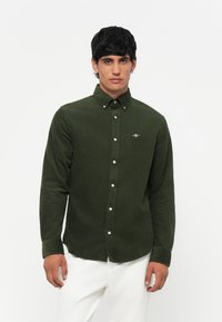 Donkergroene corduroy overhemd met een button-down kraag, witte knopen en een klein geborduurd logo op de borst. Lange mouwen en een getailleerde pasvorm.