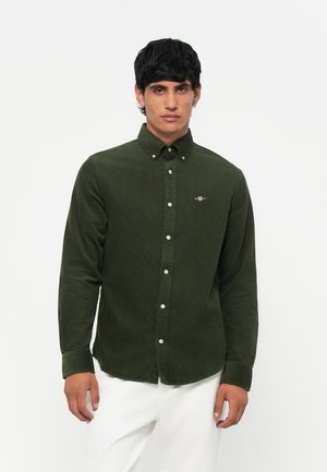 Camisa de veludo cotelê verde escuro com colarinho abotoado, botões brancos e um pequeno logótipo bordado no peito. Mangas longas e corte ajustado.