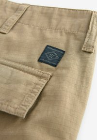 Beige stofshorts med et rutemønster, prydet med et marineblåt mærke, der står "Premium Grade Durable Goods". Indeholder en subtil lomme detalje.