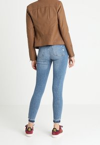 Giacca in ecopelle marrone, jeans skinny blu chiaro e sneakers borgogna. La giacca presenta una parte posteriore testurizzata e chiusure a zip basilari.