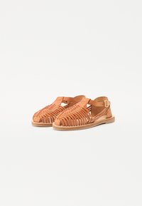 Lysbrune lærsandaler med åpen design, vevde remmer, justerbar ankelspenne og flat såle. Glatt tekstur med naturlig finish.
