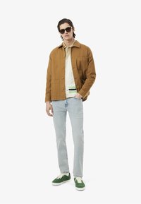 Giacca di cotone marrone sopra una camicia bianca con bottoni, abbinata a jeans blu chiaro e sneakers verdi. Accessoriata con occhiali da sole scuri.