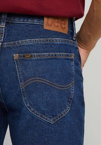 Blå denim jeans med en teksturert overflate, med et brunt skinnmerke, kontraststing og et buet baklomme-design.