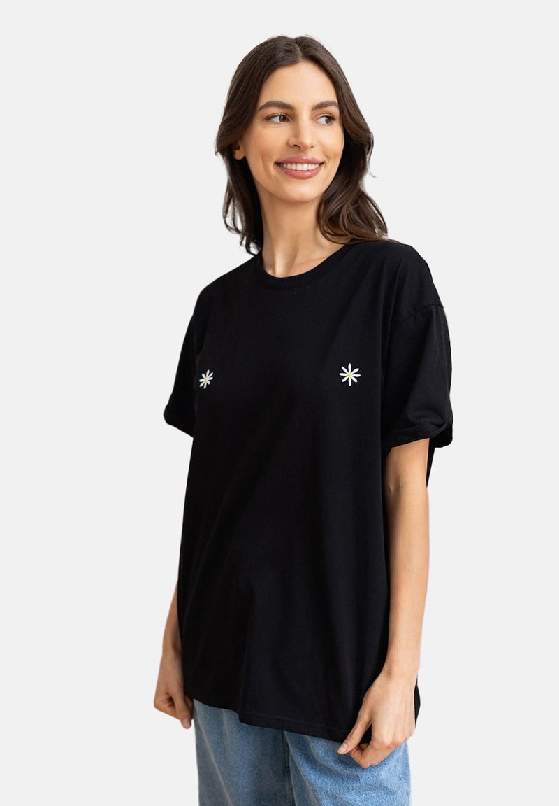 Czarna bawełniana koszulka z krótkim rękawem, z dwoma haftowanymi białymi gwiazdami na klatce piersiowej i luźnym krojem. Widoczne niebieskie dżinsy.