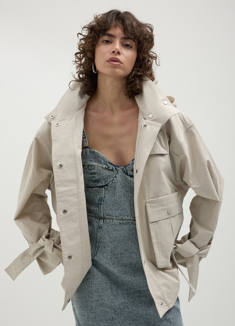 Veste beige surdimensionnée avec un col montant, des fermetures à pression et une poche avant. Fabriquée en tissu lisse avec une coupe décontractée.