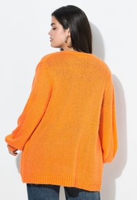 Pull en tricot orange surdimensionné avec un ourlet côtelé, des manches longues et un design texturé, présenté sur un fond blanc.