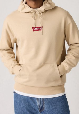 Personne portant un sweat à capuche beige Levi's avec poche avant et capuche à cordon, associé à un jean bleu et un sous-vêtement blanc visible sous l'ourlet.