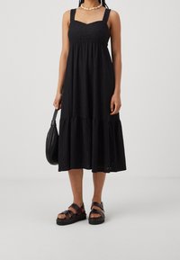 Schwarzes Baumwollkleid mit quadratischem Ausschnitt, breiten Trägern und gestuftem Saum. Mit Lochstickerei und fällt bis zur Mitte der Waden. Getragen mit dunklen Sandalen.