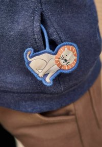 Tengerészkék szövet, bézs és narancs árnyalatú oroszlán felvarróval, hímzett részletekkel, ami egy zseb közelében van rögzítve.
