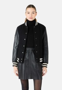 Zwarte varsityjas met een wollen lichaam en leren mouwen, voorzien van witgestreepte ribgebreide manchetten en een voorste drukknoopsluiting. Gecombineerd met een zwarte rok.