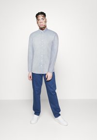 Cămașă lungă cu mânecă bleu deschis, cu butoane, asociată cu pantaloni negri și adidași albi, prezentată pe un fundal alb simplu.
