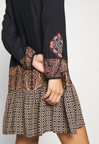 Schwarzes Kleid mit langen Ärmeln, das über eine aufwendige florale und Paisley-Stickerei in Rot, Grün und Beige verfügt, abgeschlossen mit einem gerafften Saum.