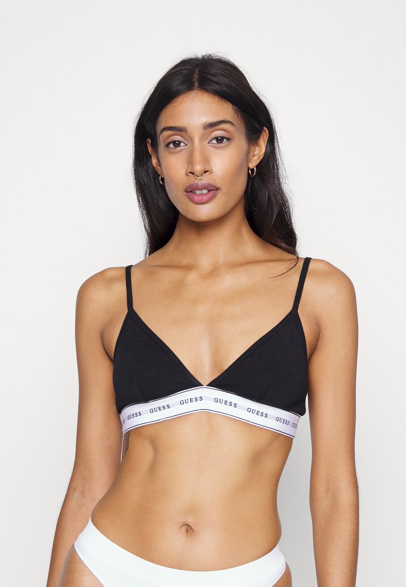 Black cotton bralette with thin straps, triangular cups, and a white band featuring "GUESS" in purple lettering. Smooth texture and casual design.