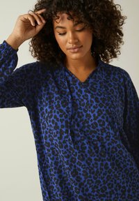 Blå leopardmönstrad blus med V-ringning, långa ärmar och elastiska manschetter. Tyget verkar slätt och lätt, med djärva svarta detaljer.